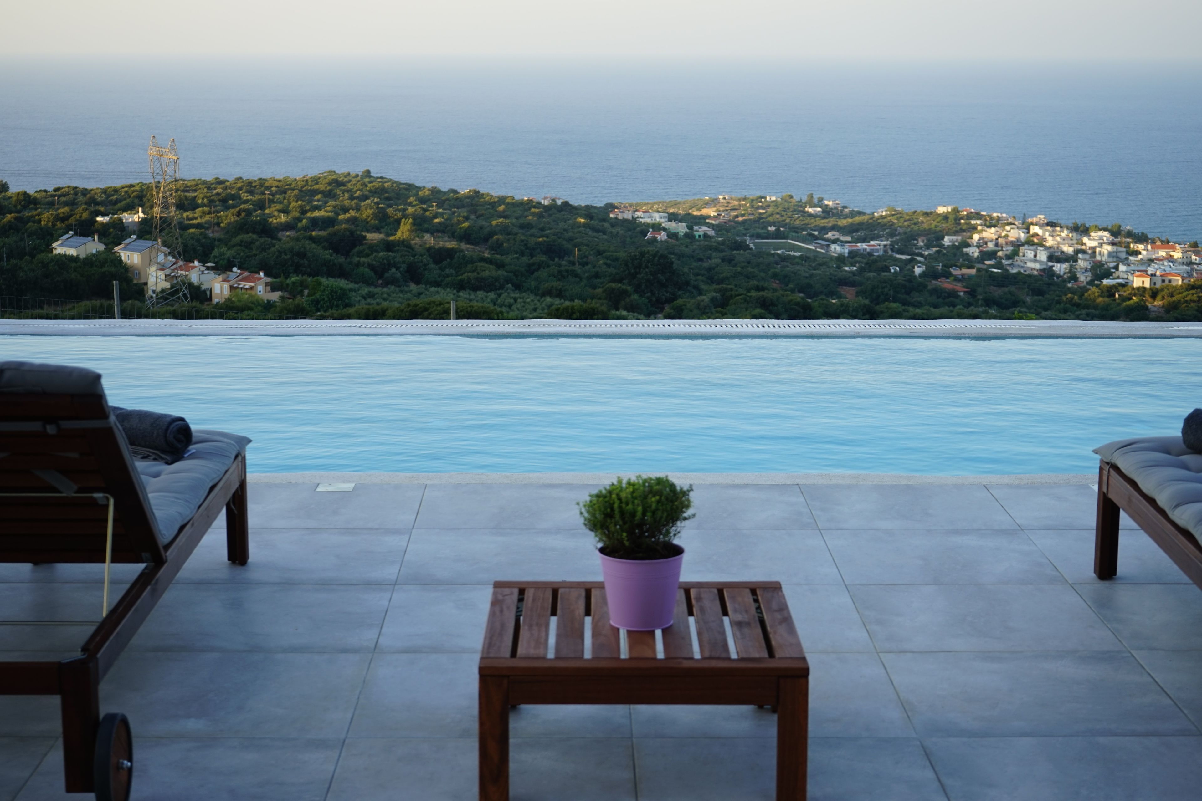 A poolside area with two wooden lounge chairs, a small table, and a potted plant. The background features a view of a coastal town and the sea.
