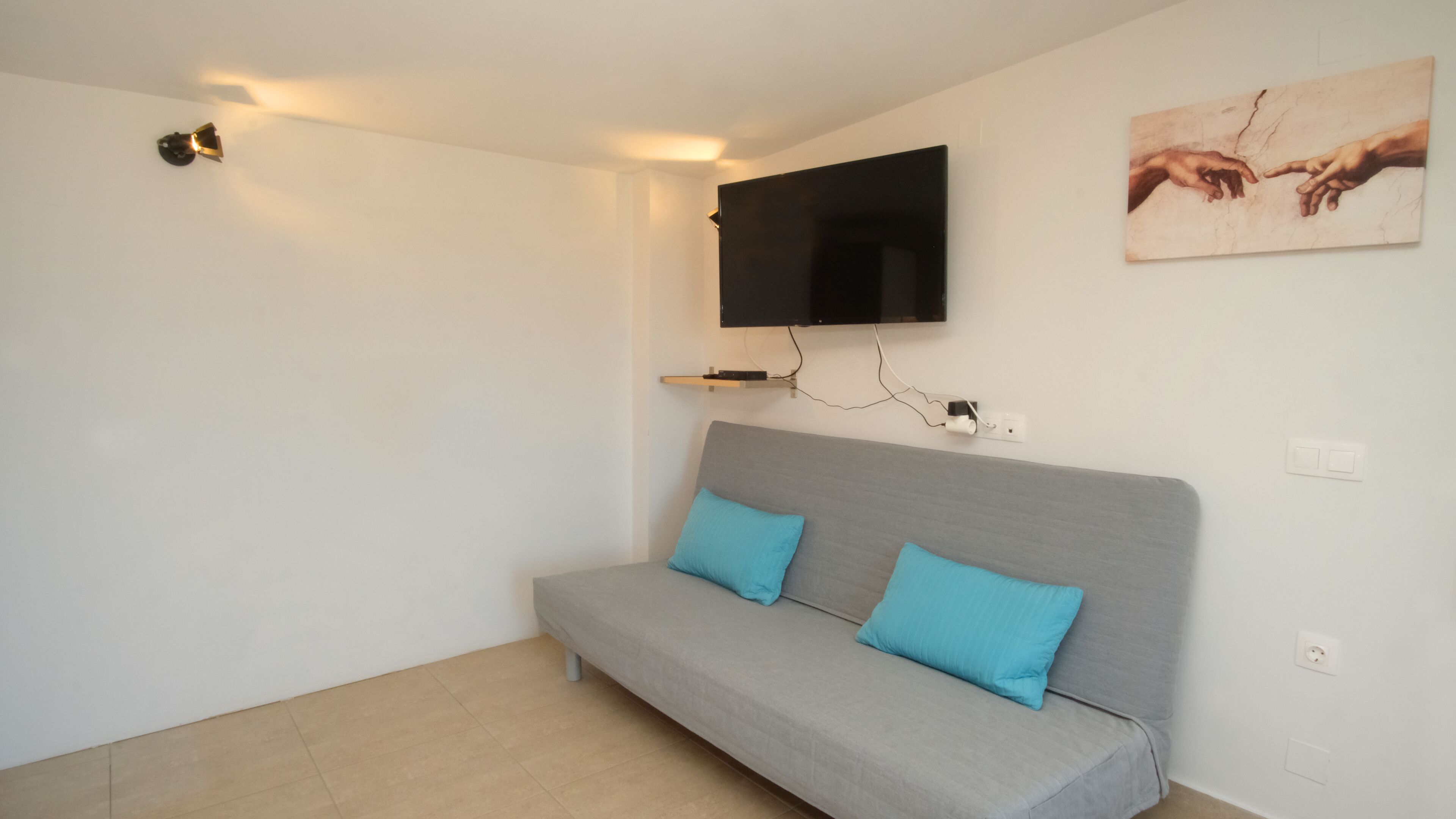 Een moderne woonkamer met een grijze bank, twee blauwe kussens, een wandgemonteerde flatscreen televisie en een plank aan de muur.