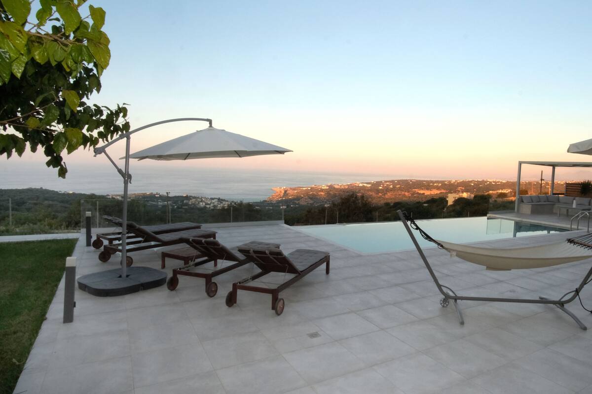 A rooftop pool area with lounge chairs, umbrellas, and a view of the cityscape at sunset.