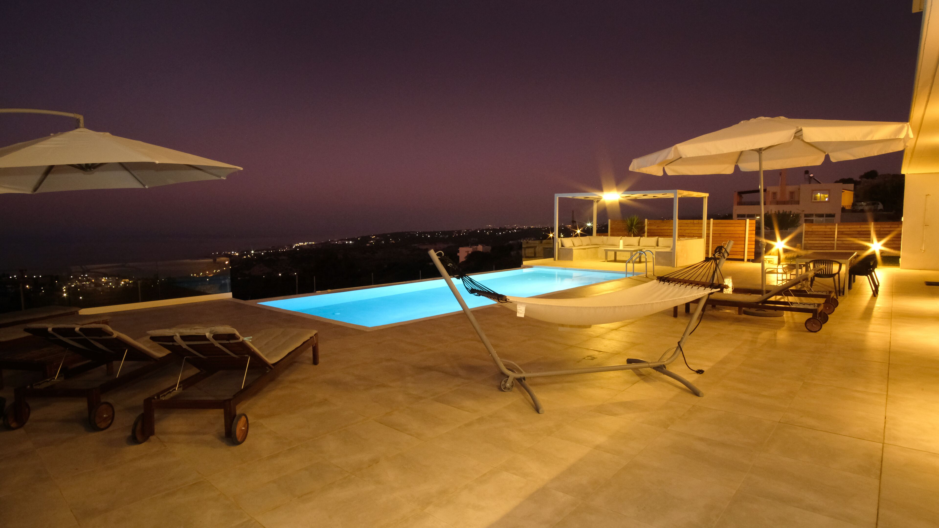 A rooftop pool area with lounge chairs, umbrellas, and a view of the city at night.
