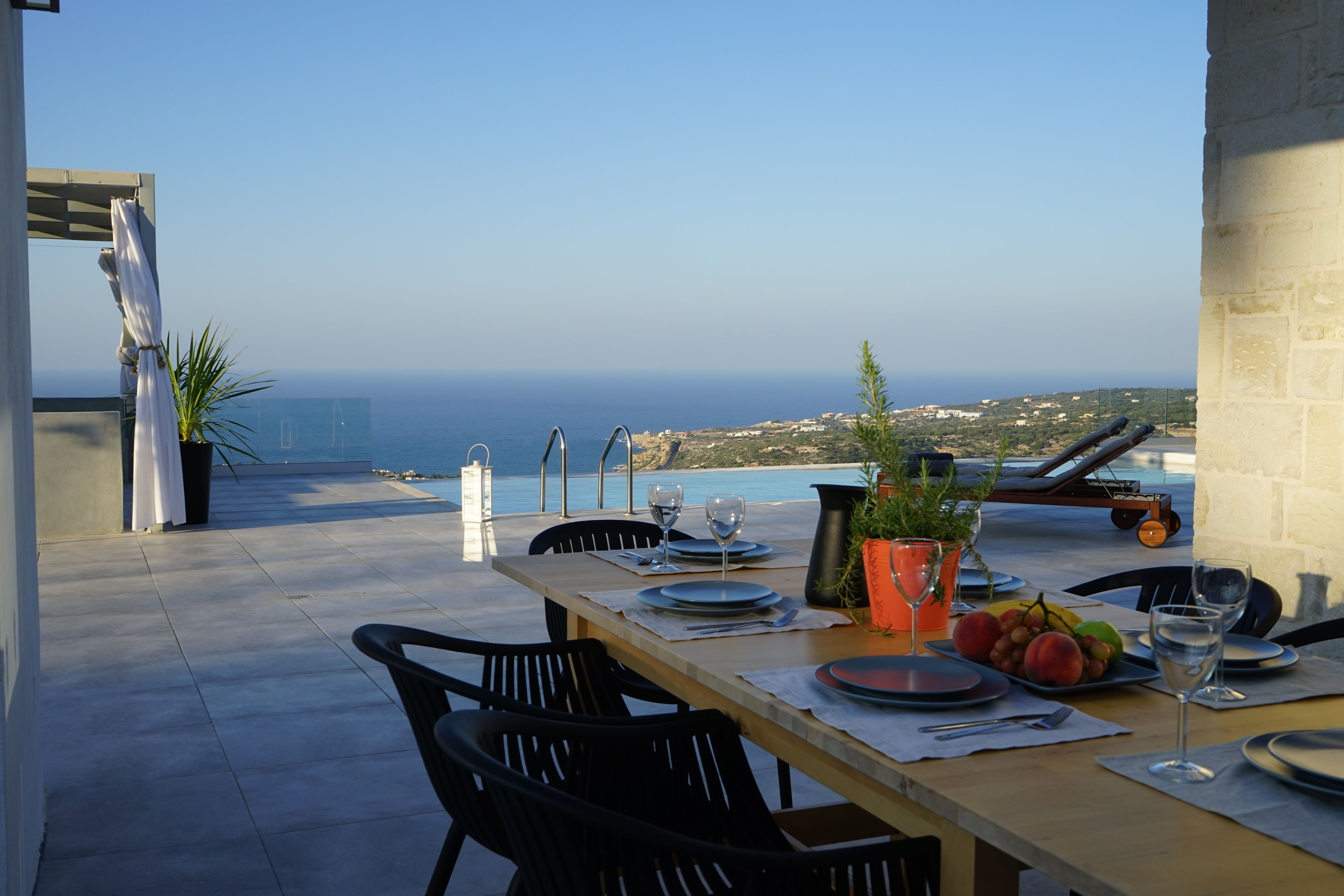An outdoor dining area with a table set for two, overlooking a scenic view of the sea and distant landscape.