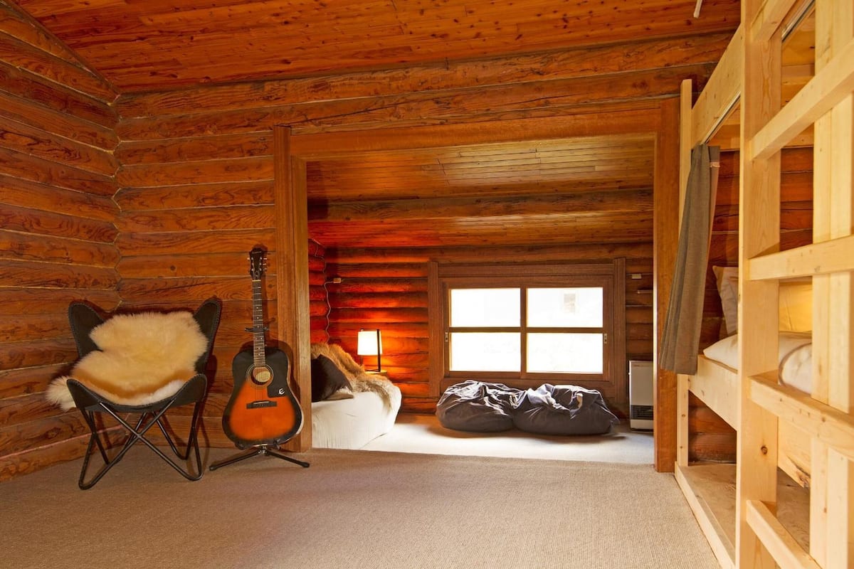 Interior de una cabaña de troncos con una guitarra, un sillón de piel, un sillón de plumas y una cama litera.