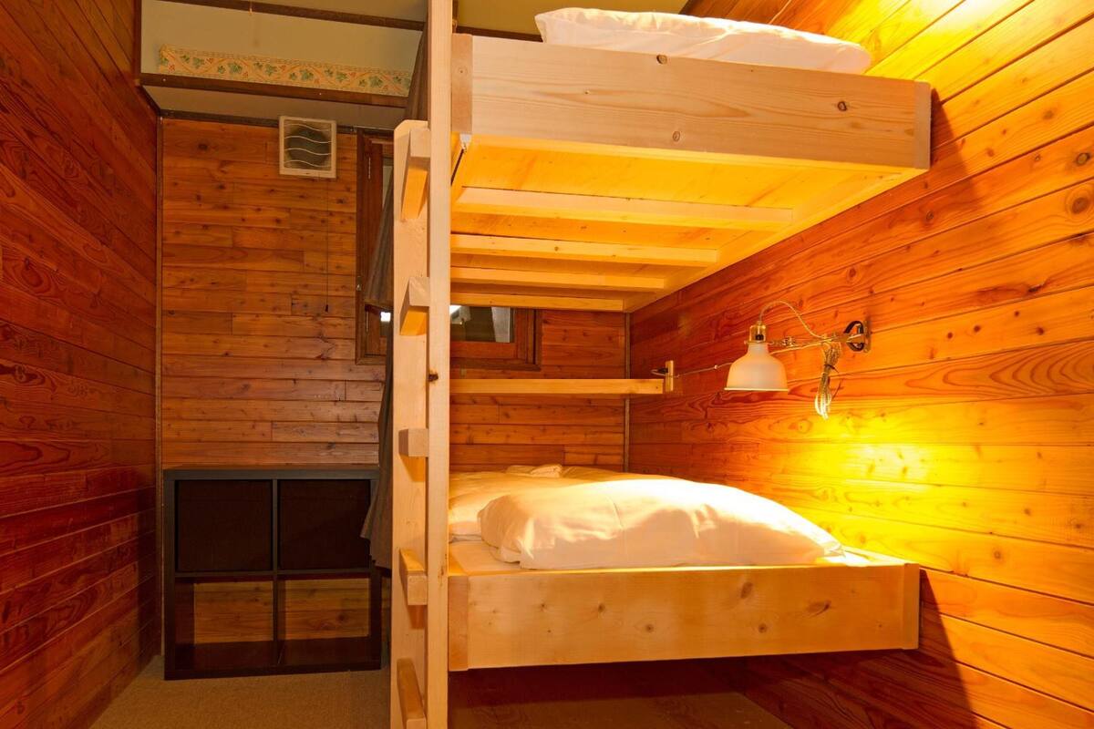 Una habitación tipo cabaña de madera con una cama nido, un ventanal pequeño y una lámpara fijada a la pared.