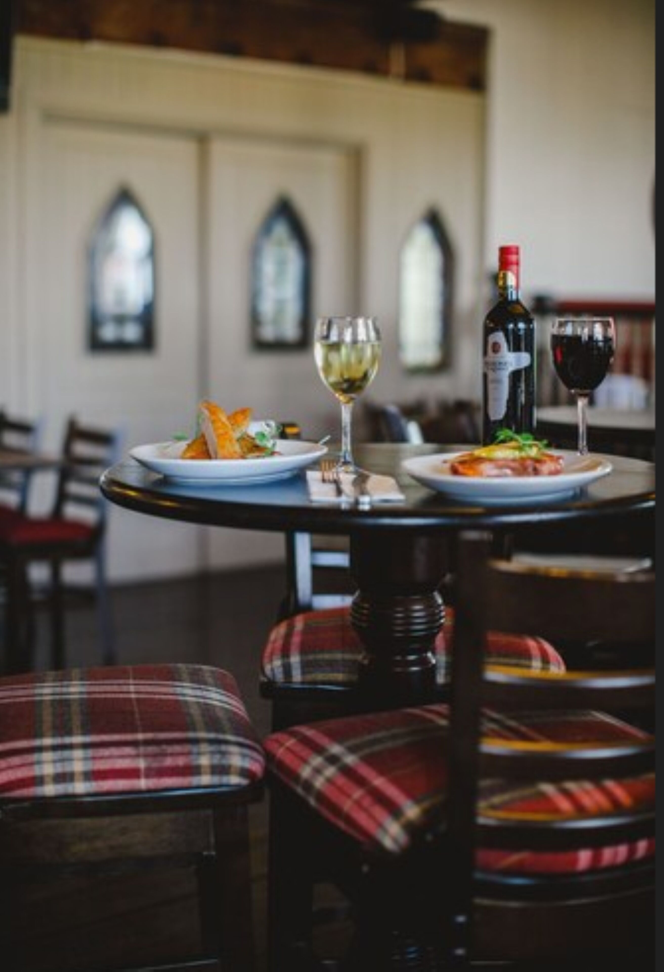 A dining table with plates of food and glasses of wine, set in a room with plaid chairs and arched windows.