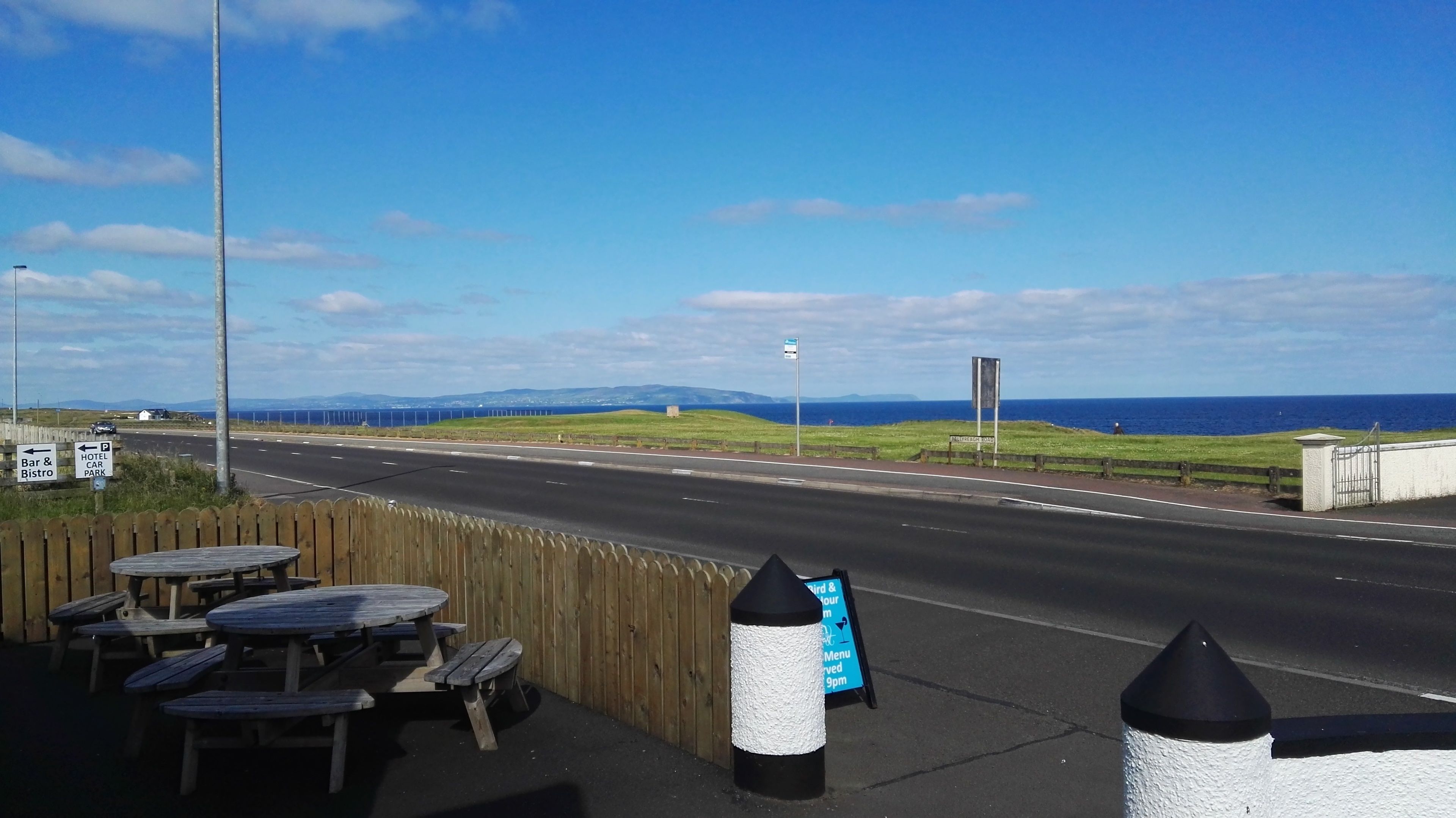 A coastal road with a view of the sea, picnic tables, and directional signs for a hotel and car park.