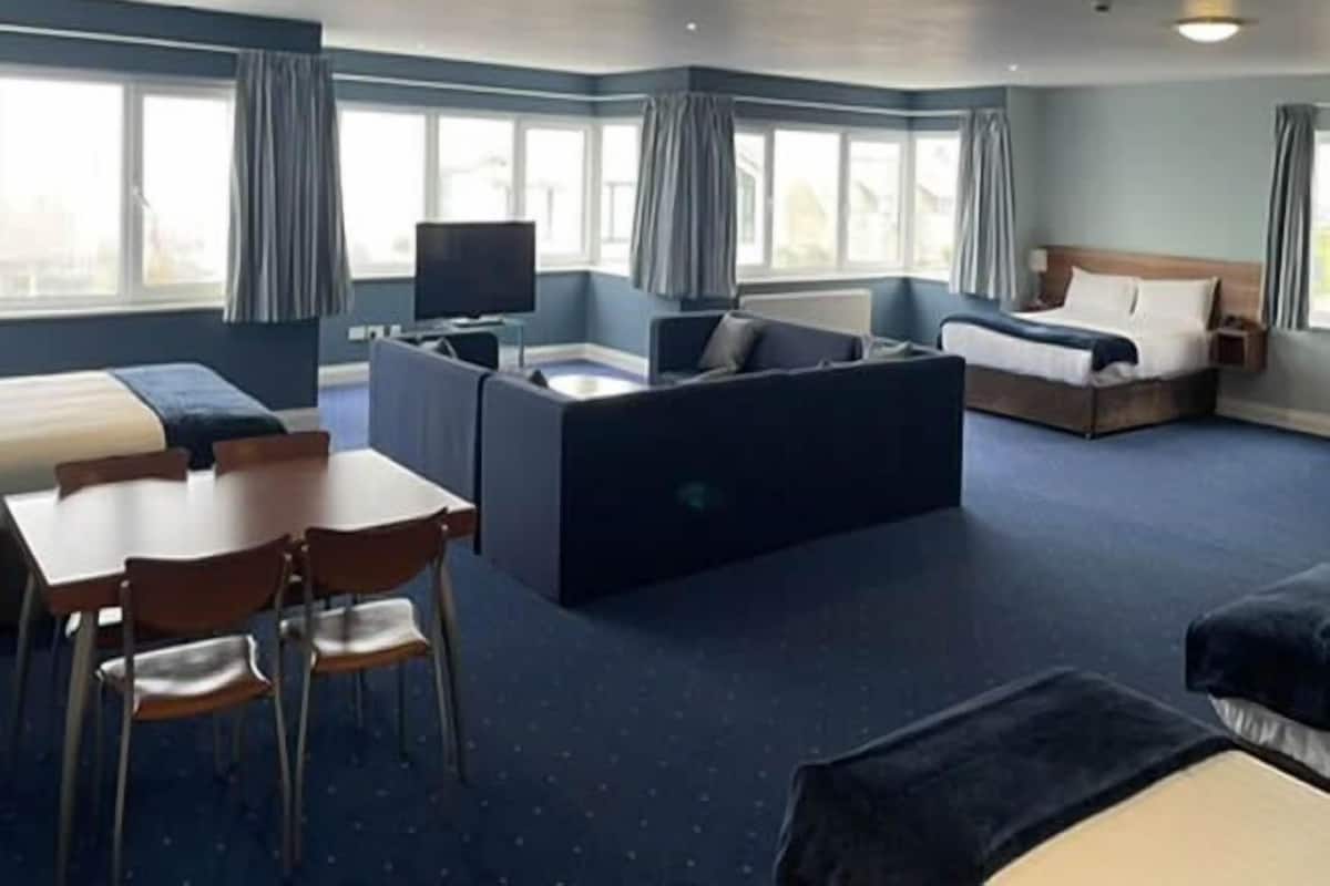 A hotel room with a bed, a sofa, a desk with chairs, a television, and large windows.