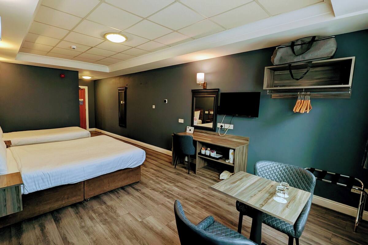 A hotel room with a double bed, a desk with a chair, a TV, and a small table with two glasses.