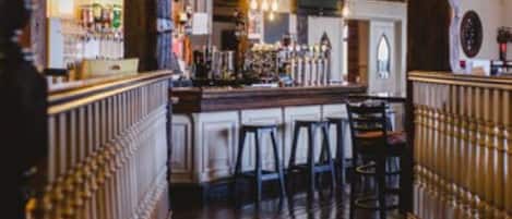 A bar area with wooden beams, a bar counter, stools, and a kitchen in the background.