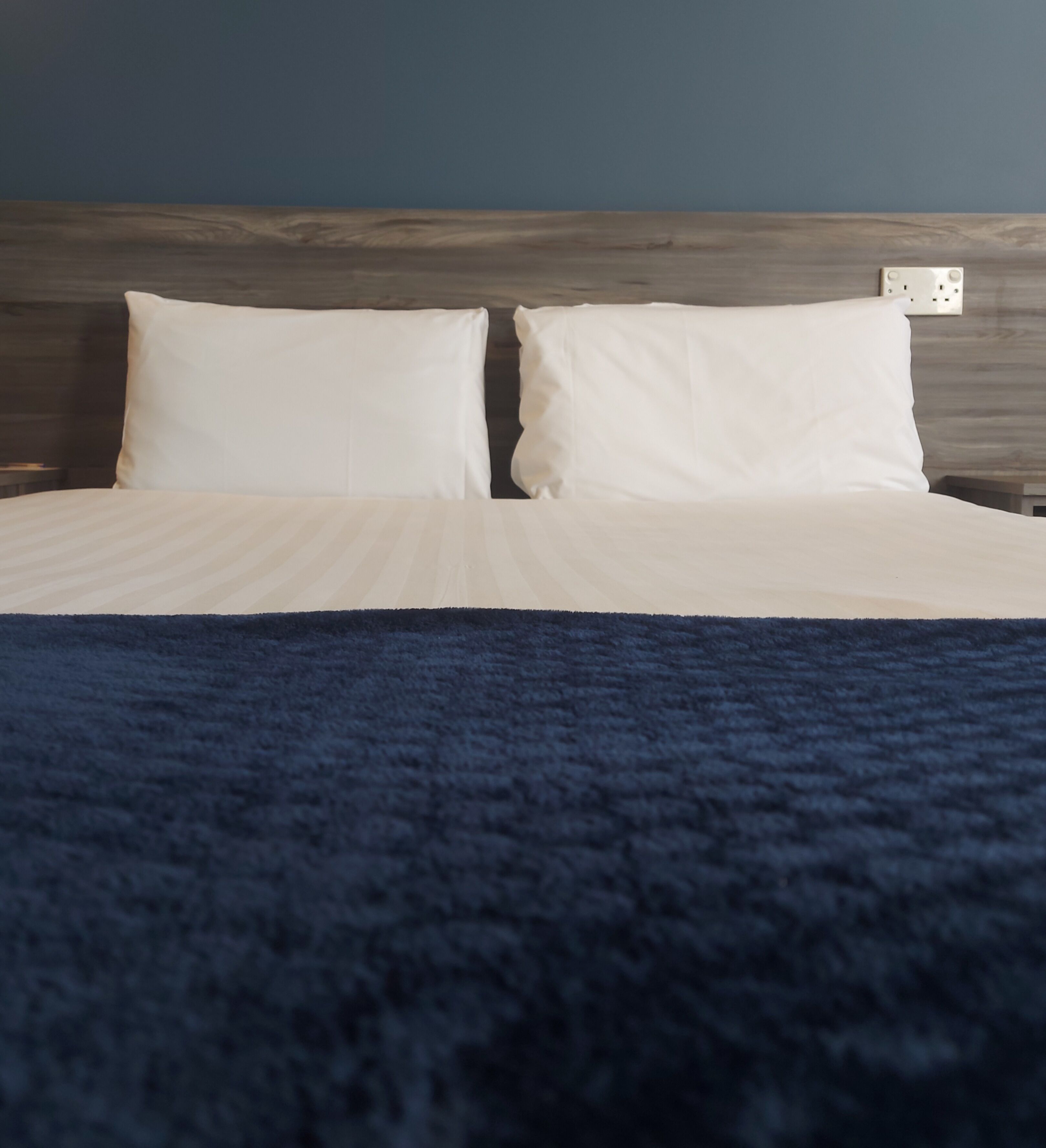 A bed with white linens and a dark blue blanket, a wooden headboard, and a wall socket.
