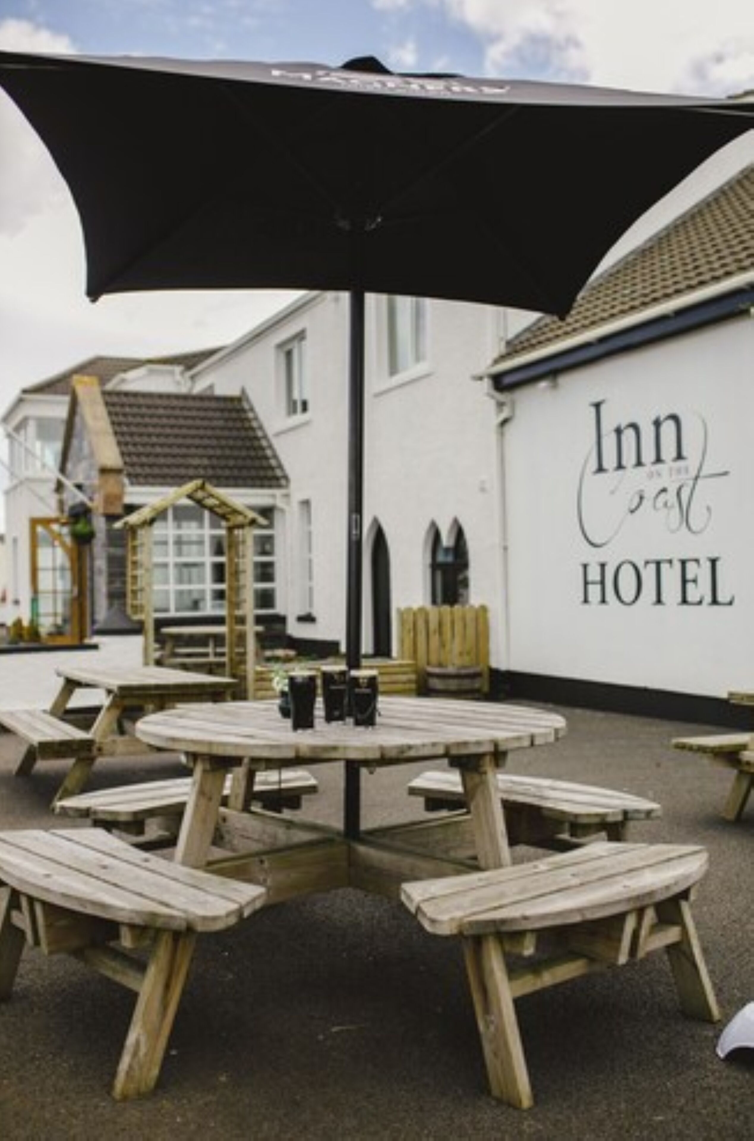 Et træbænkebord med bænke og en sort parasol foran en hvid bygning med skiltet "Inn on the coast HOTEL".