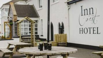 Et træbænkebord med bænke og en sort parasol foran en hvid bygning med skiltet "Inn on the coast HOTEL".
