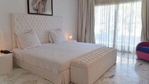 Una cama grande con cabecera capitonada, una mesita de noche con lámpara y una ventana con cortinas translúcidas.