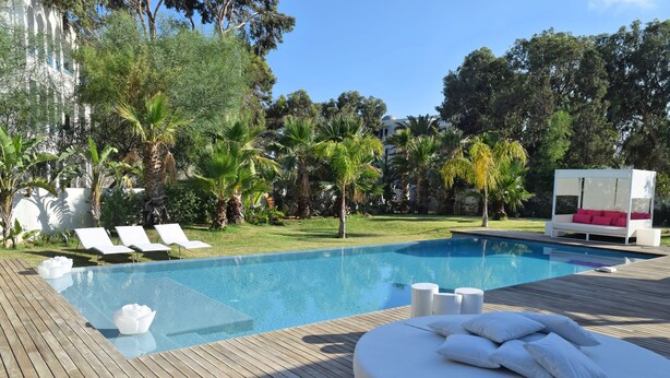 Zona de piscina con terraza de madera, sillas de descanso y una cabaña blanca con cojines rojos.