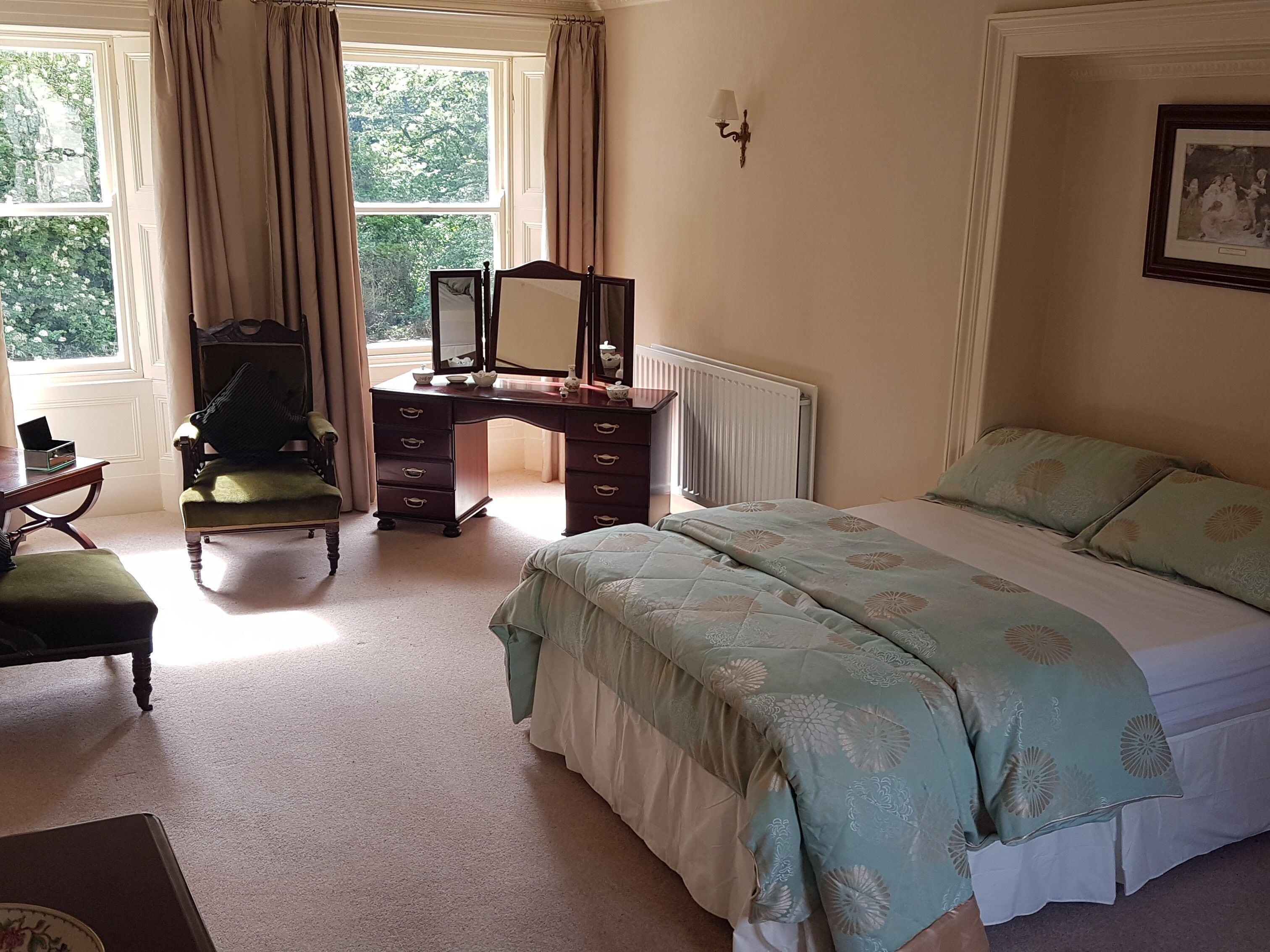 A bedroom with a bed, a desk with a chair, a radiator, and a window with curtains.