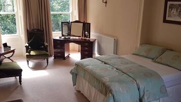 A bedroom with a bed, a desk with a chair, a radiator, and a window with curtains.