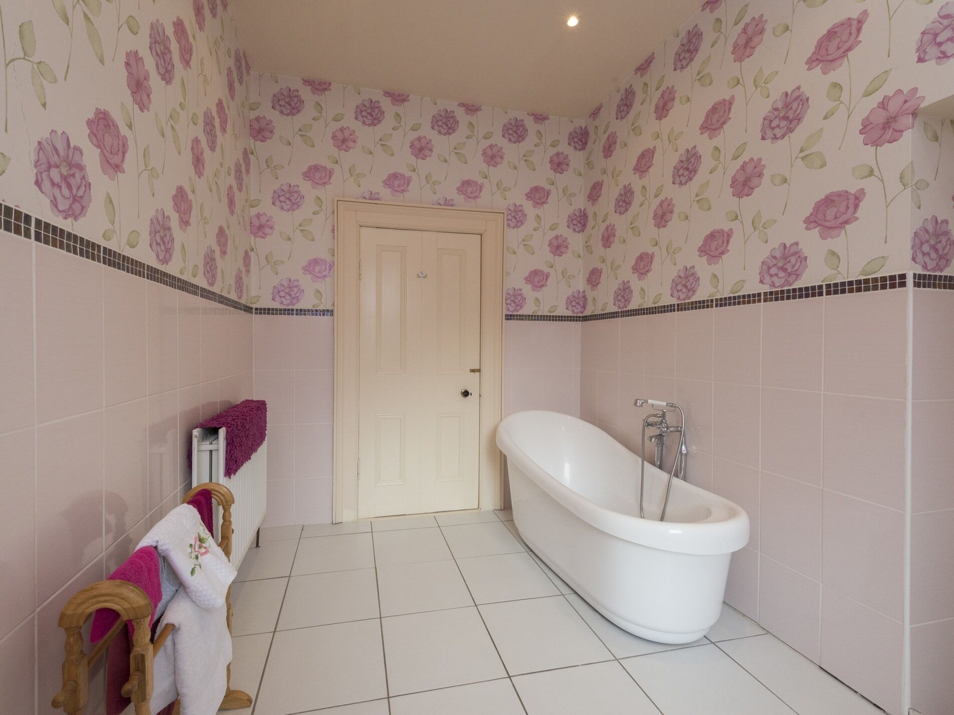 Un baño con una bañera independiente, un sillón de madera y una toalla rosa, con papel tapiz floral.