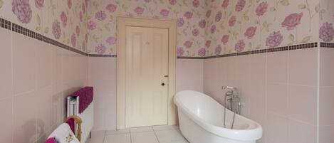 Un baño con una bañera independiente, un sillón de madera y una toalla rosa, con papel tapiz floral.