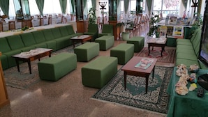 Uma ampla área de estar com sofás, pufes e mesas verdes, com um tapete estampado e uma televisão.