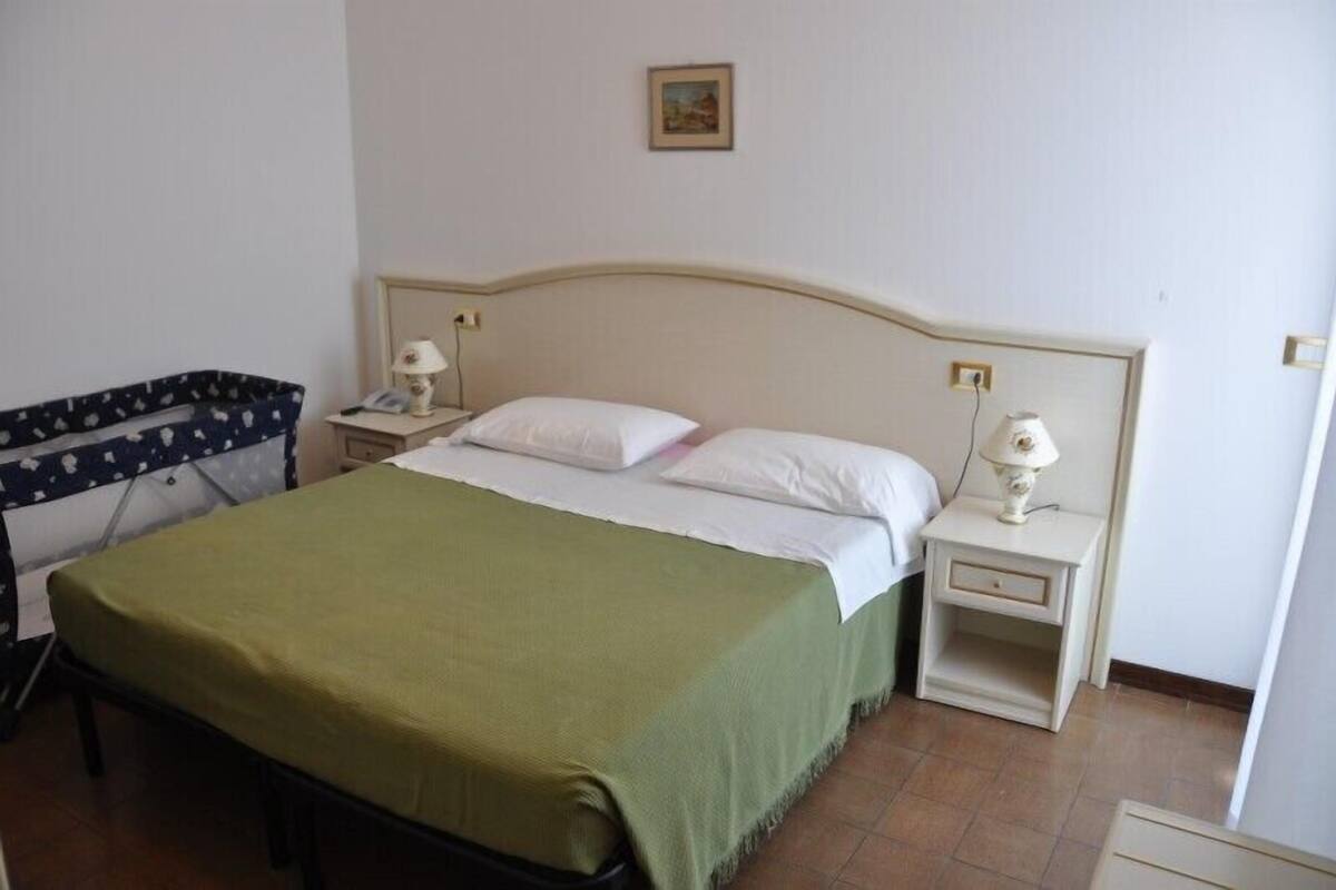 Habitación con una cama individual, colcha verde, dos mesitas de noche con lámparas y una mesita auxiliar con lámpara, además de un cuadro en la pared.
