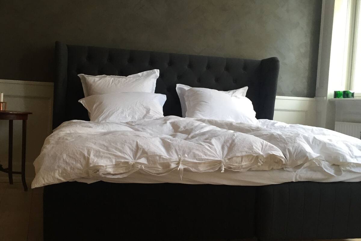 Un dormitorio con cabecera oscura, suelo de madera y mesita de noche con lámpara.