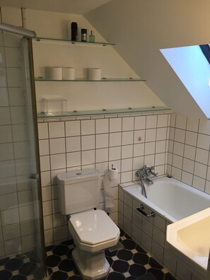 Un baño con inodoro, lavamanos y bañera. Hay estantes sobre el inodoro y un tragaluz sobre la bañera.