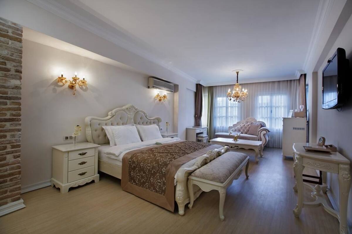 A spacious bedroom with a large bed, a sitting area with a sofa and armchair, a small table, and a television mounted on the wall.