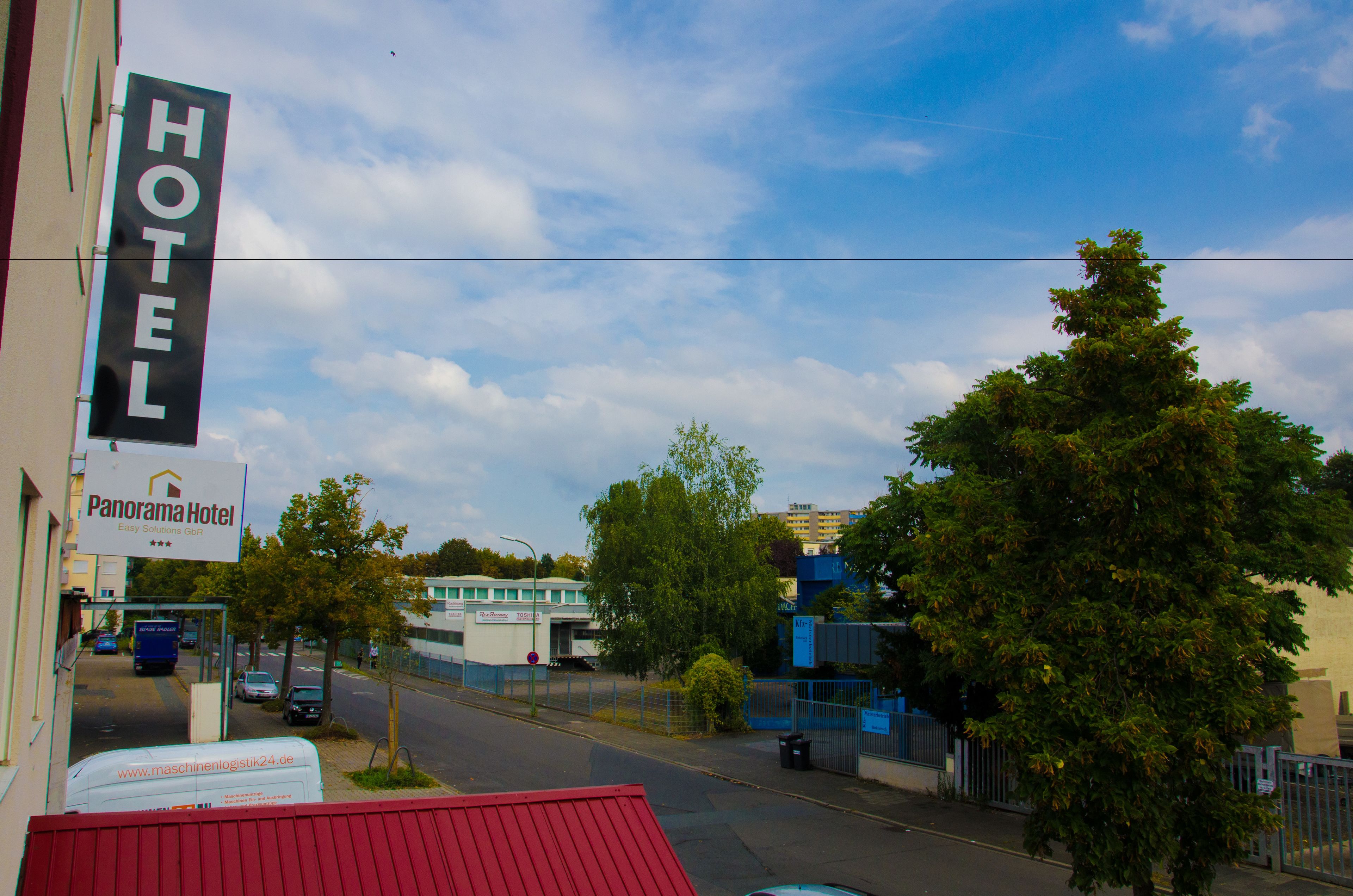 A hotel sign with "HOTEL" and "Panorama Hotel" text, a street view with a blue building, trees, and a fence.