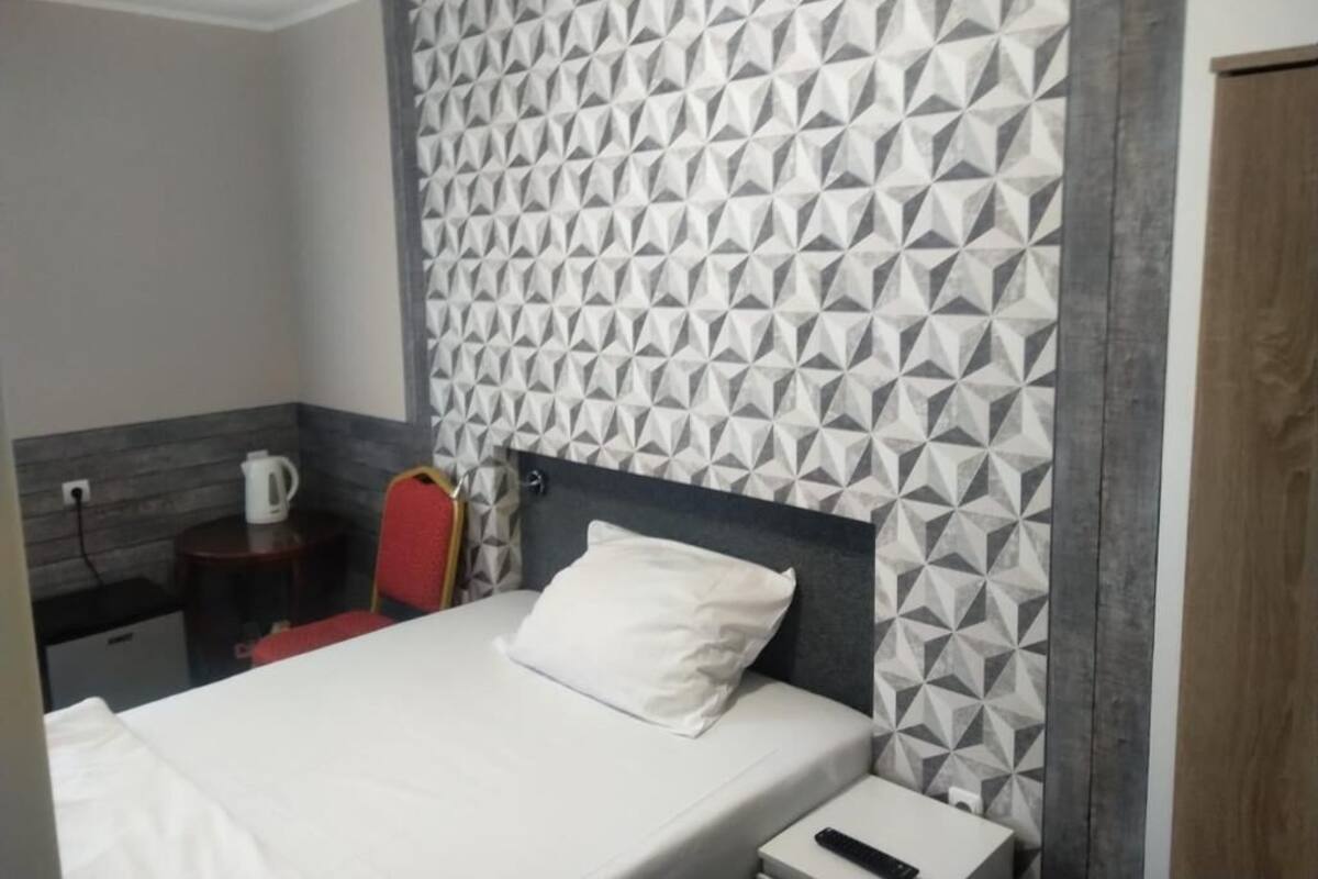 A hotel room with a geometric-patterned wall, a bed with a white pillow, a bedside table with a remote control, and a red chair.