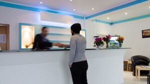 A reception area with a counter, a seated guest chair, and a small table with a vase of flowers. - Panorama Hotel (Offenbach am Main)