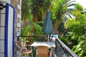 A balcony with a table and two wicker chairs, surrounded by lush tropical plants and a pool in the background. - Anaxos Garden (Lesvos)
