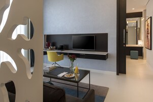 Una sala moderna con una televisión de pantalla plana montada en la pared, una cafetera, una computadora portátil y una mesa de comedor con un jarrón de flores.