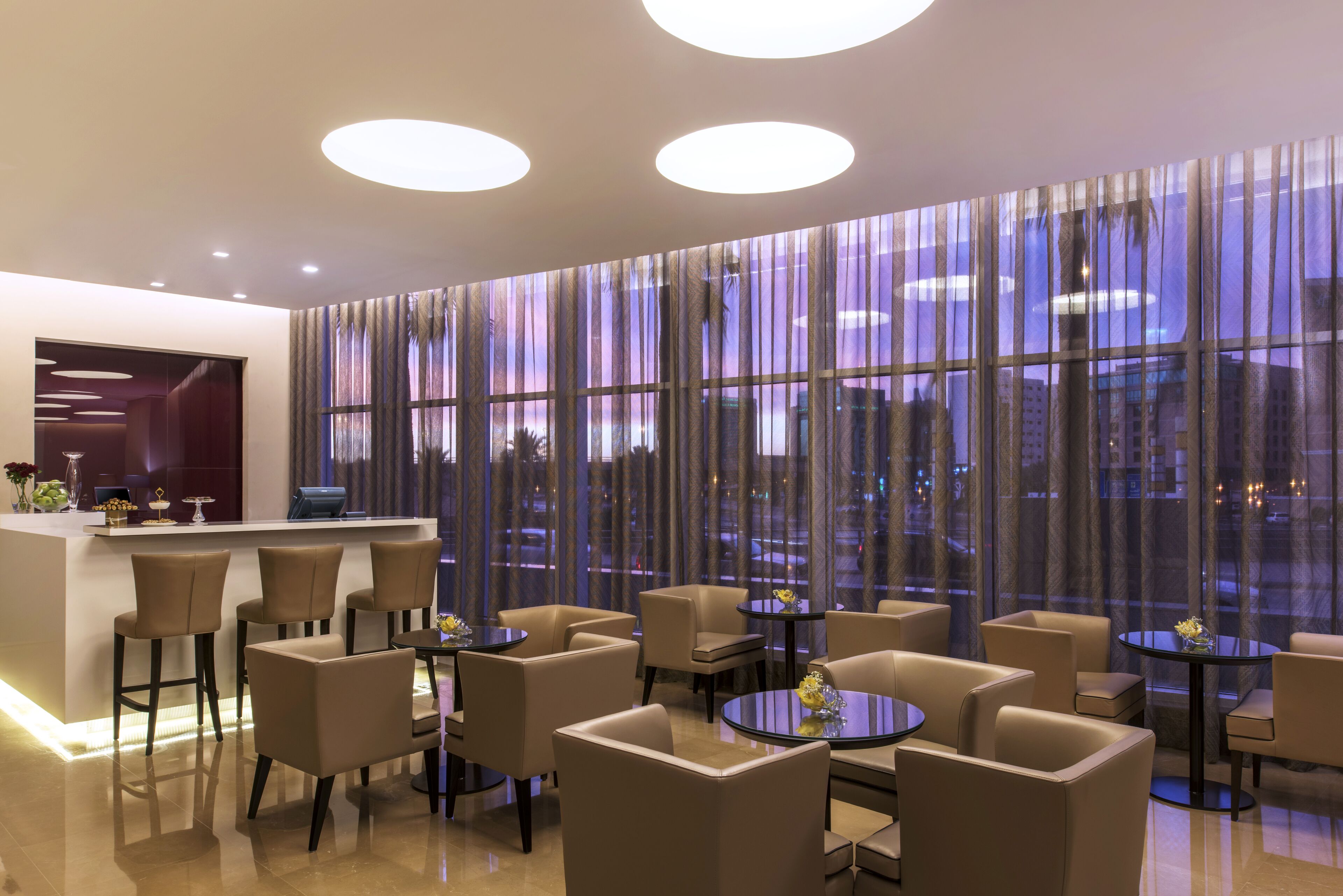 Un moderno vestíbulo de hotel con una recepción, varias zonas de estar y grandes ventanales con cortinas.