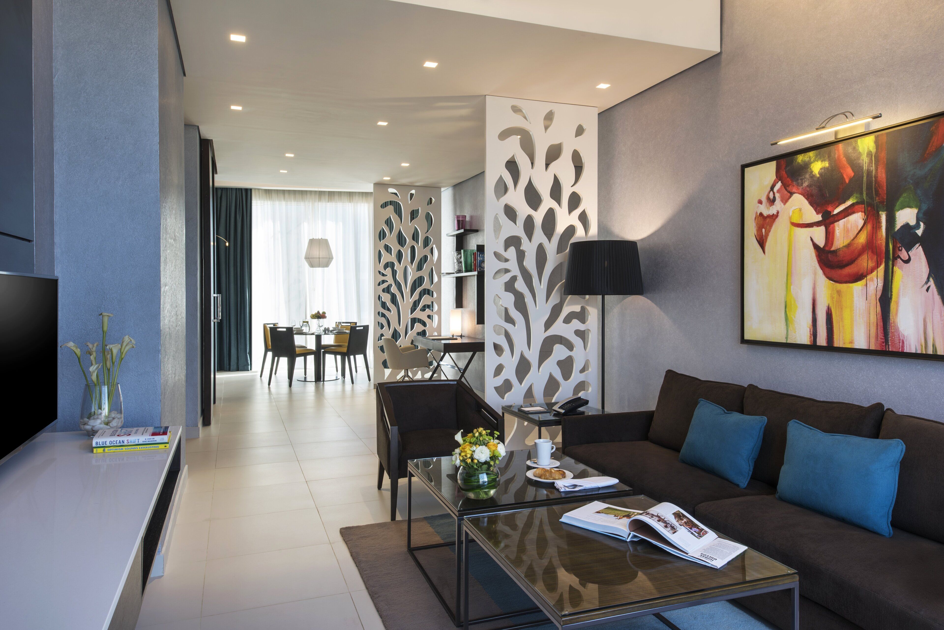 A modern living room with a glass coffee table, a brown sofa with blue cushions, a TV on the left, and a dining area in the background.