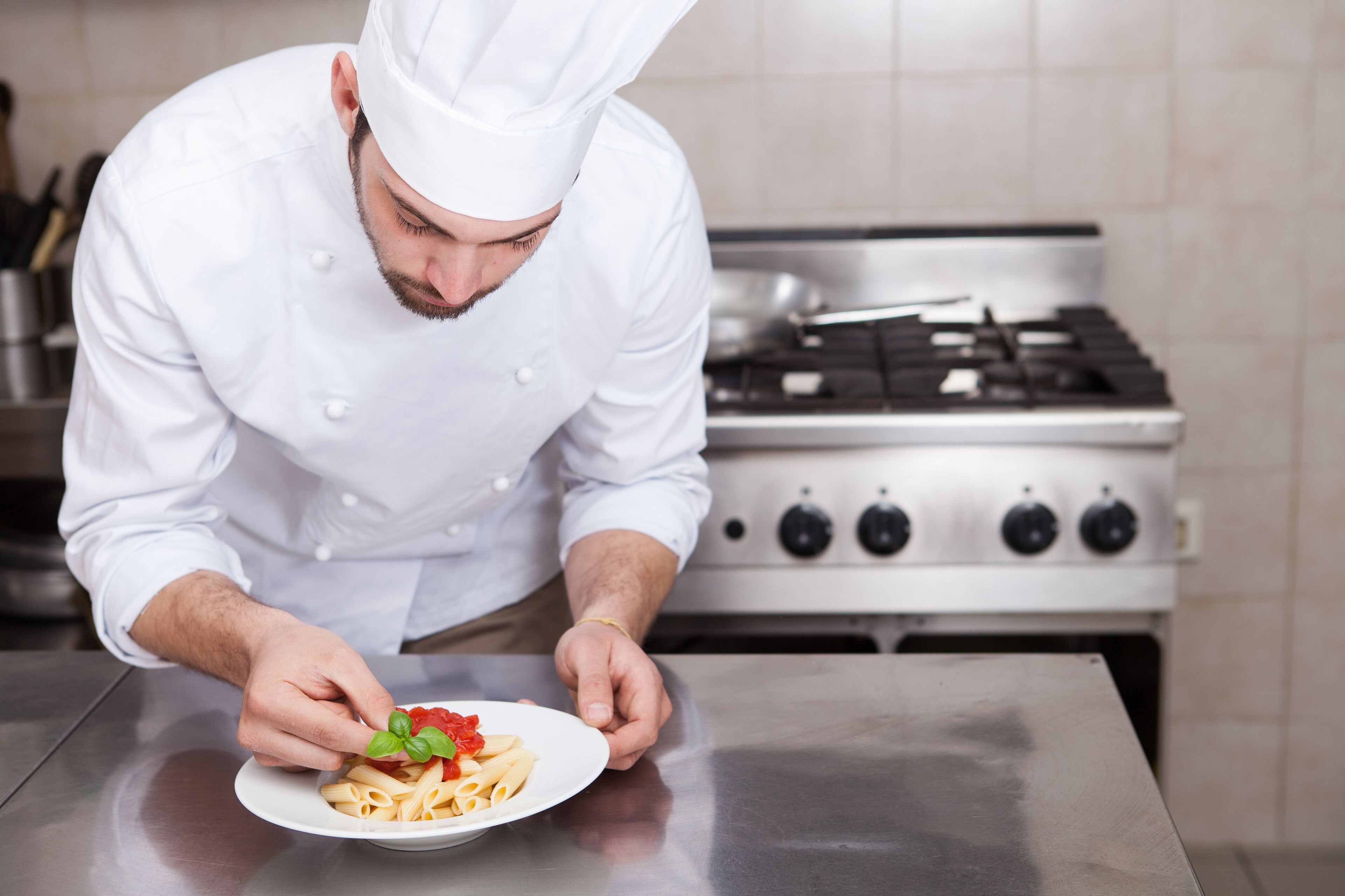 Um chef com uniforme branco preparando um prato com massa e folha de manjericão.