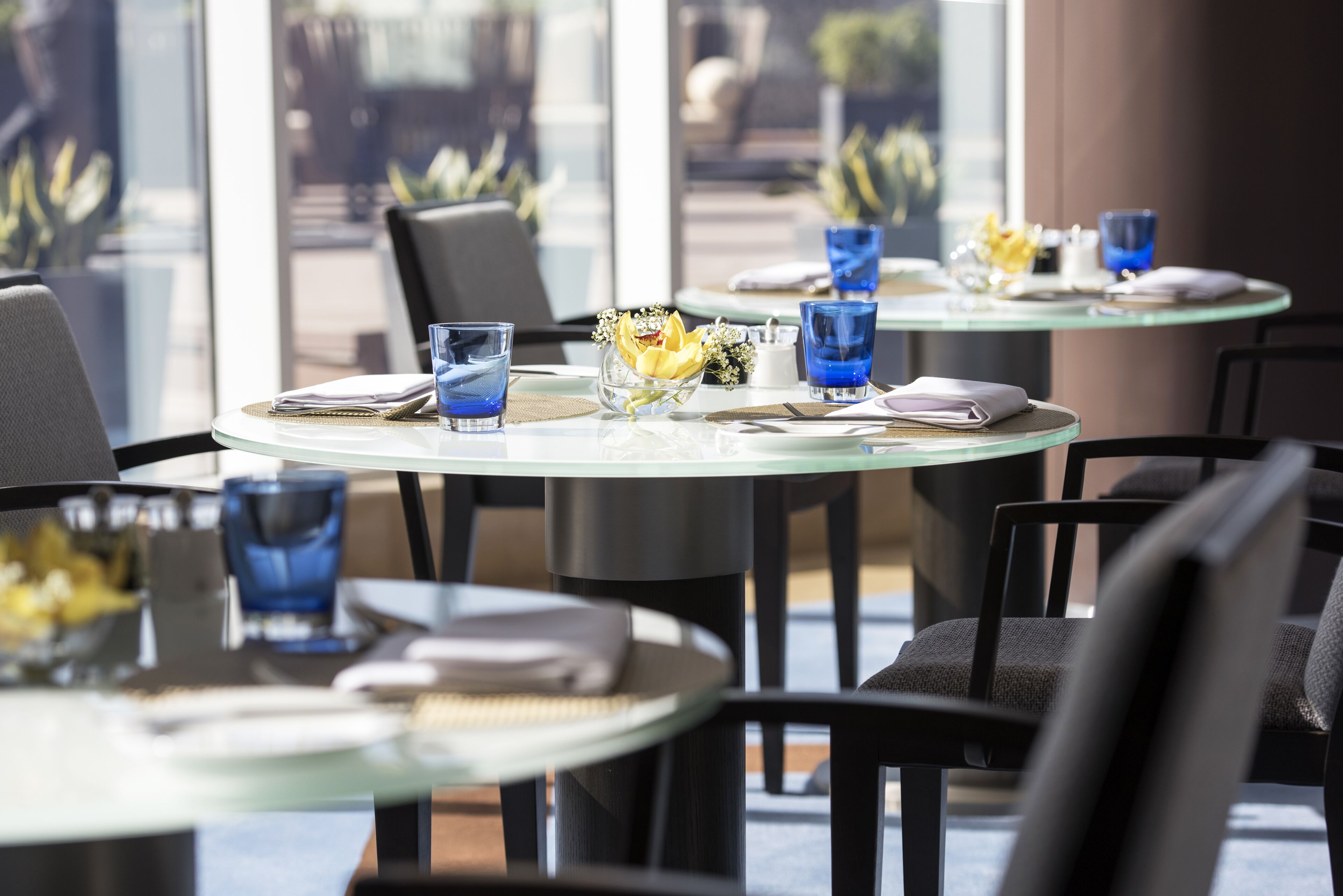 Un restaurant avec des tables rondes en verre, de la verrerie bleue et des fleurs jaunes disposées sur les tables.