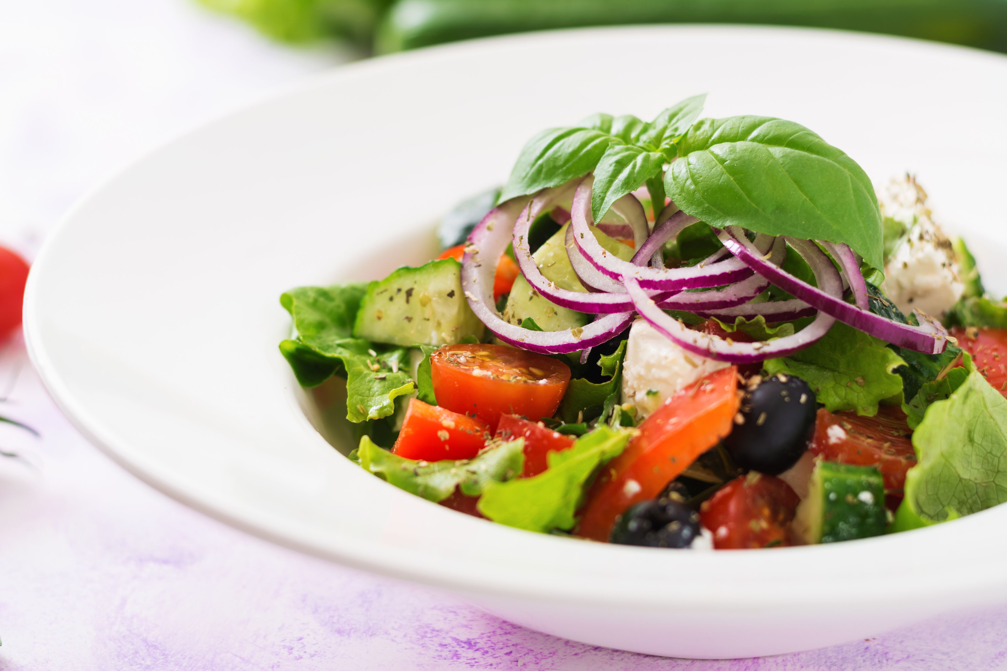 A plate of mixed greens salad with tomatoes, cucumbers, and red onions, garnished with a basil leaf.