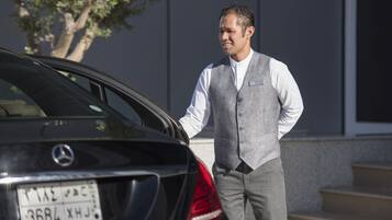 A man in a grey vest and white shirt standing next to a black car with a visible license plate.