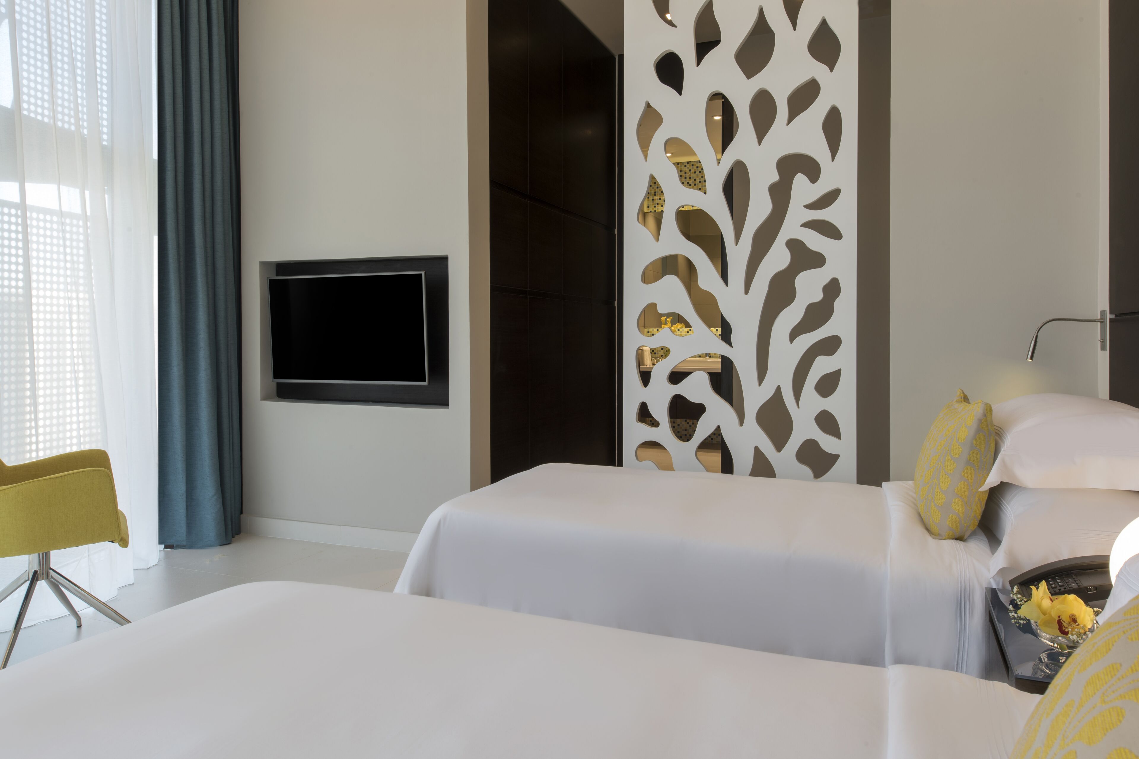 Una habitación de hotel moderna con dos camas, un televisor montado en la pared, un separador decorativo y una silla amarilla.