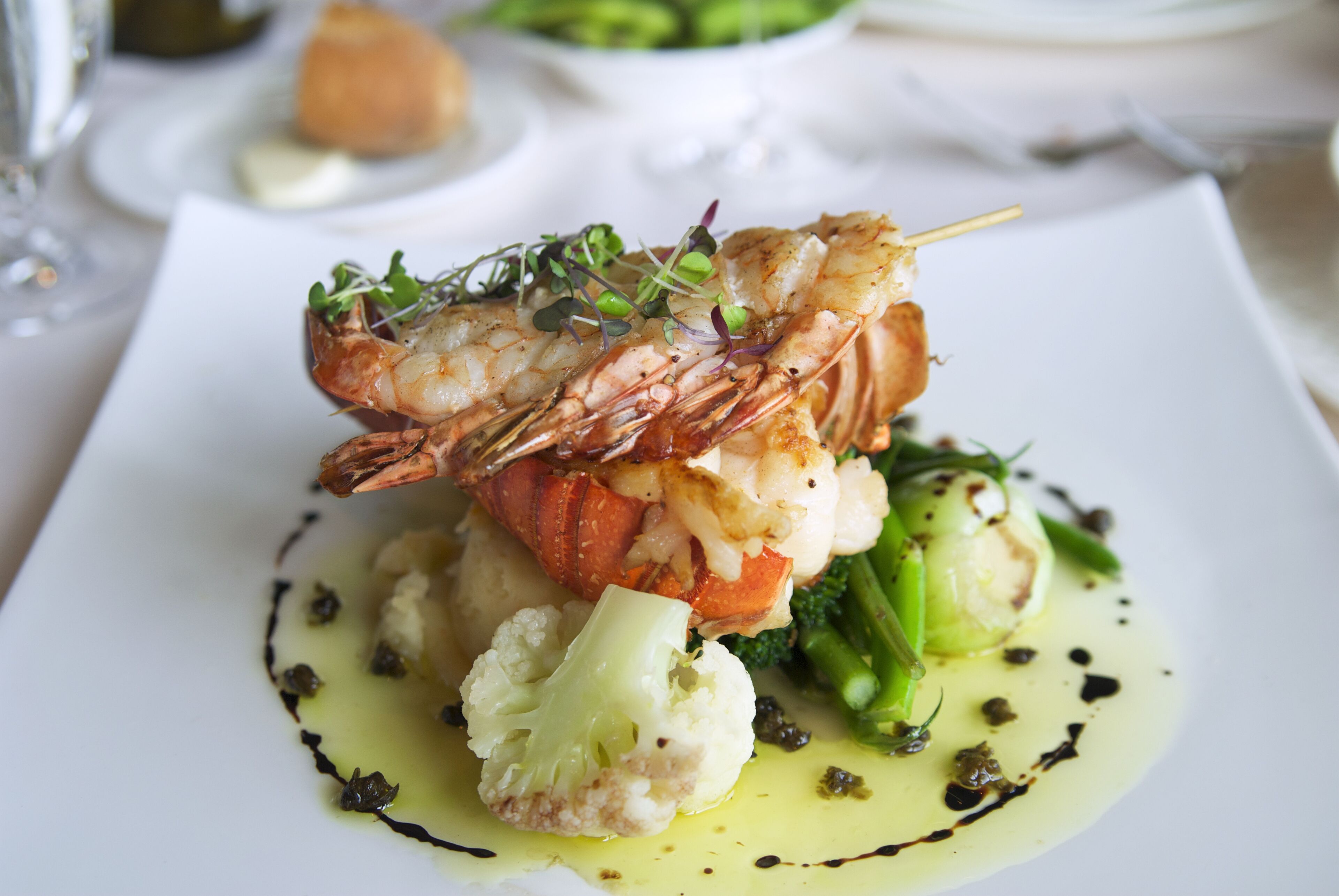 A seafood dish with a lobster tail, cauliflower, and green vegetables on a white plate.