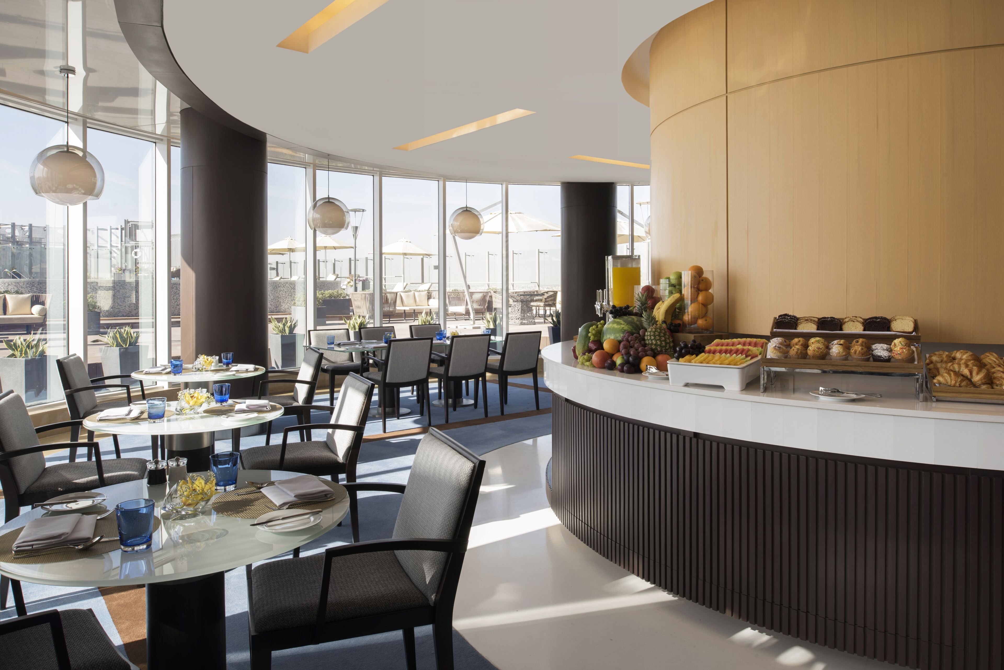 En moderne hotellobby med runde borde og stole, en central disk med frisk frugt og kager, og store vinduer med udsigt over byen.