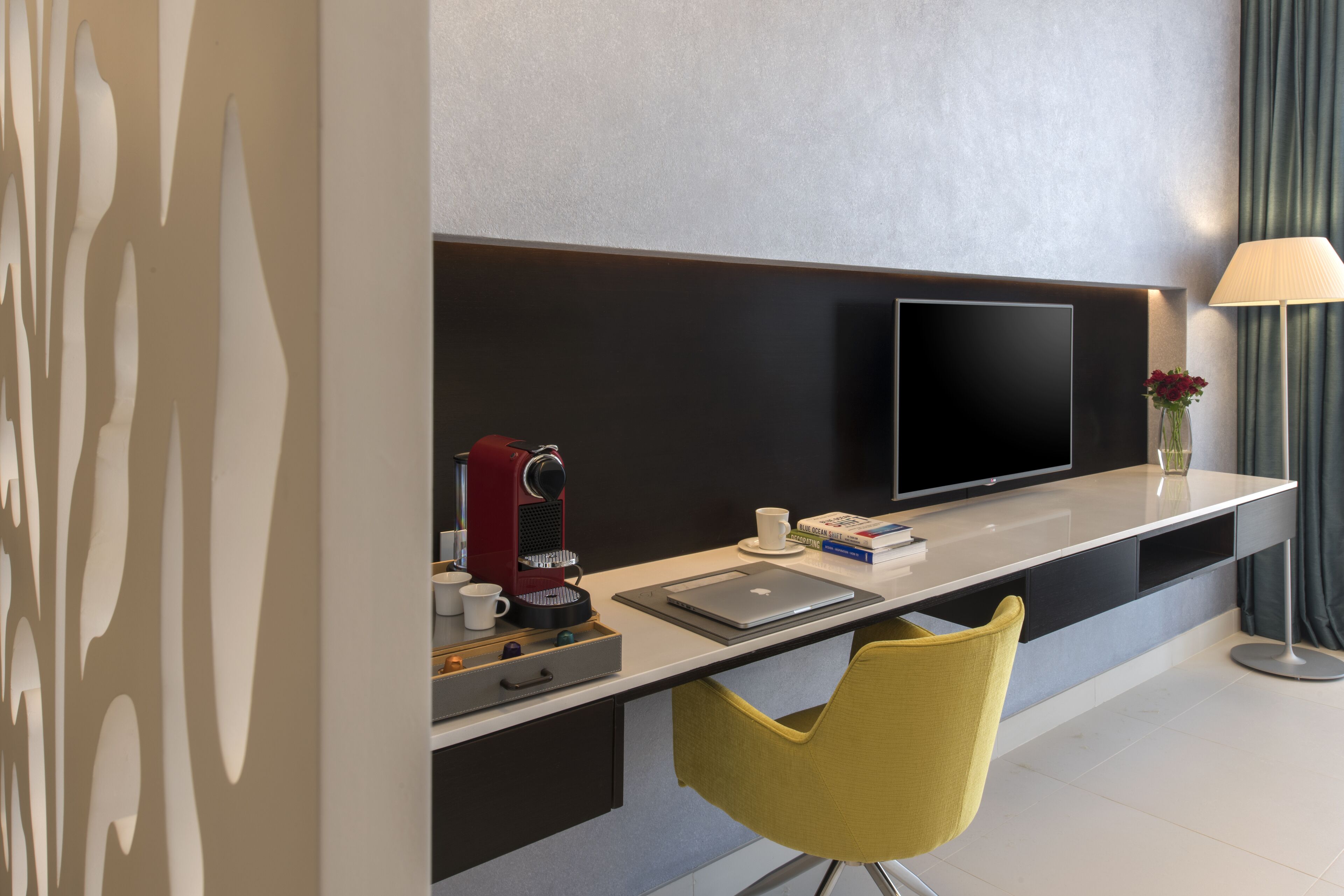 A modern hotel room with a flat-screen TV, a coffee machine, a laptop, and a yellow chair.