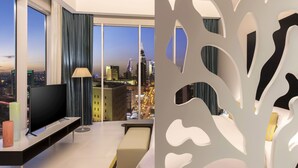 Ein modernes Hotelzimmer mit großem Fernseher, einer Sitzecke, einem kleinen Tisch und Blick auf die Skyline der Stadt im Abendrot.