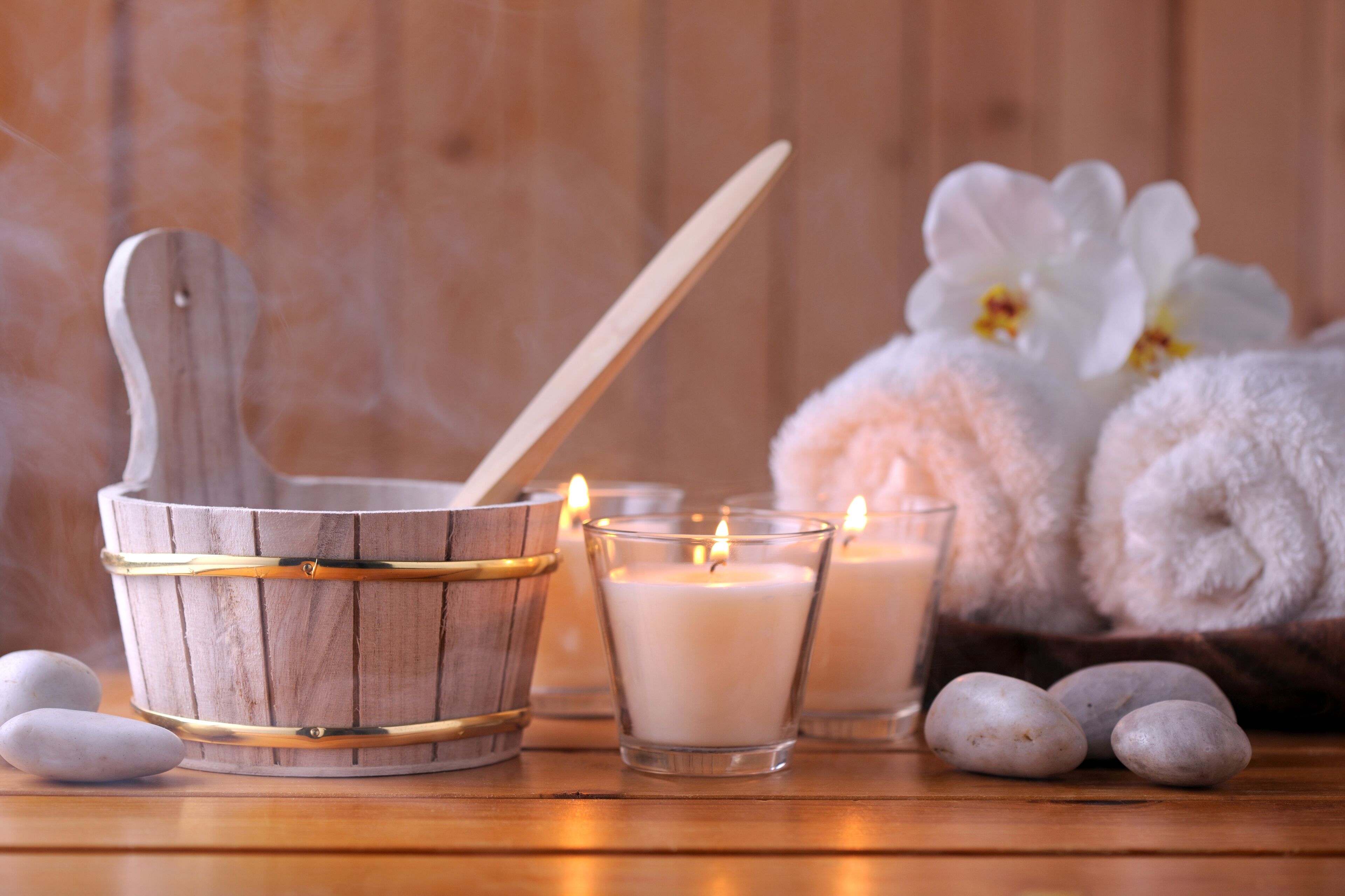 A wooden bucket with a spoon, two lit candles, and rolled towels with white flowers in the background.