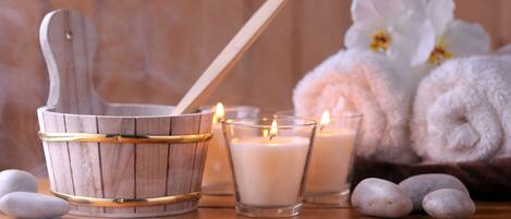 A wooden bucket with a spoon, two lit candles, and rolled towels with white flowers in the background.