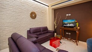 Una sala de estar con una pared de piedra, un sofá morado, un mueble bajo de madera con un televisor y un jarrón con flores.
