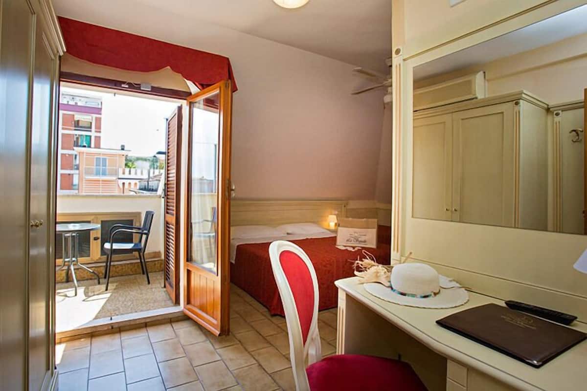 A room with a bed, a desk with a chair, a window, and a door leading to an outdoor area.