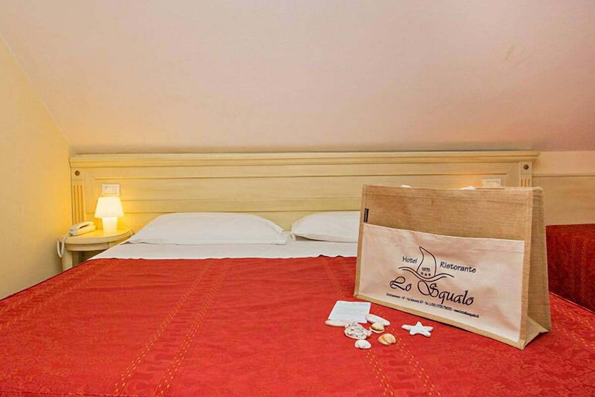 A hotel room with a bed, a nightstand with a lamp, and a bag with the hotel name on it.