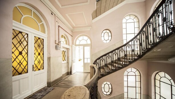 A grand staircase with ornate railings, leading to a landing with large windows and a door with yellow-tinted glass panels. - Matei Corvin Apartments (Cluj-Napoca)