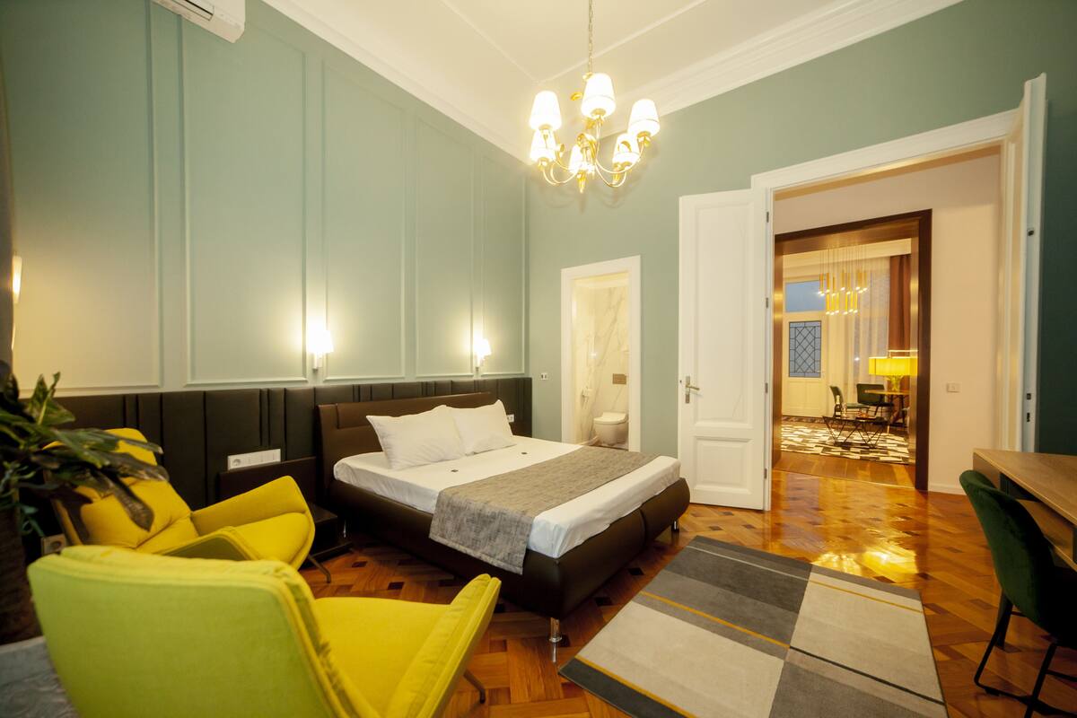 A modern hotel room with a bed, yellow chairs, a chandelier, and a view into the dining area.