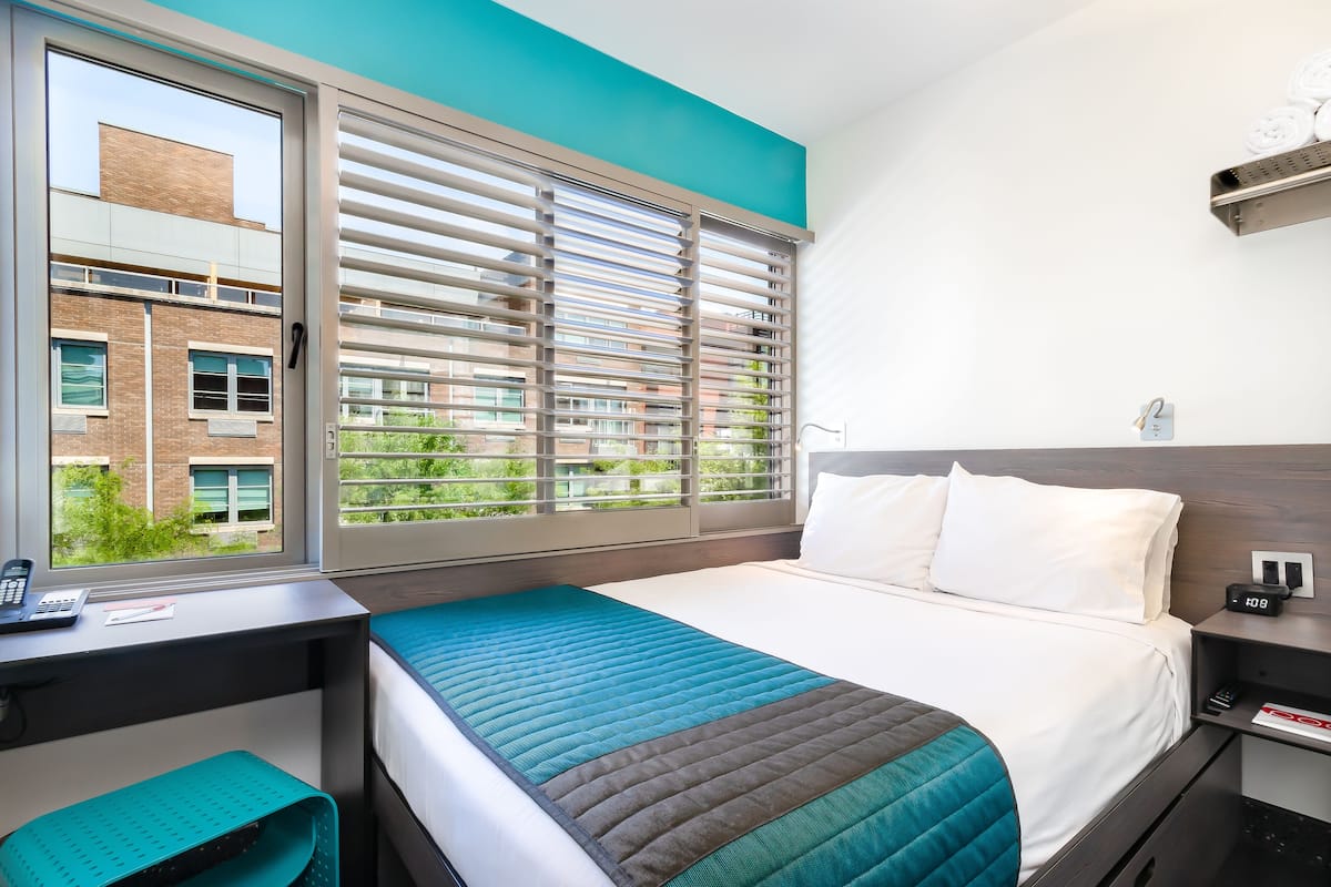 A modern hotel room with a large window, a desk, and a bed with a teal and brown quilt.